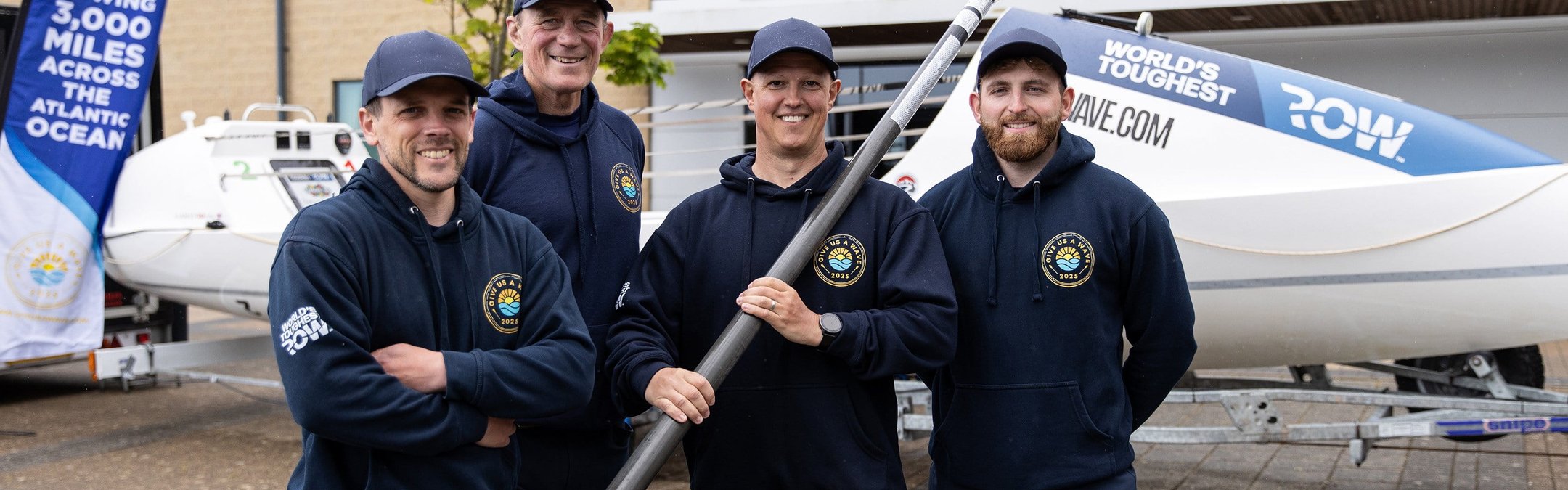 4 men standing in front of a boat. The centre right man is holding a boat paddle. They are all wearing caps and 'Give us a wave' branded hoodies.