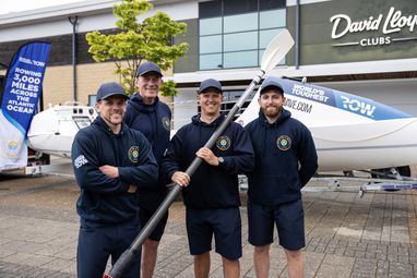 4 men standing in front of a boat. The centre right man is holding a boat paddle. They are all wearing caps and 'Give us a wave' branded hoodies.
