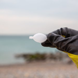 A gloved yellow hand holding an empty single-use plastic fish-shaped soy sauce container. In the background is a beach