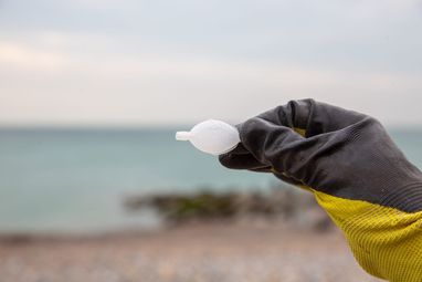 A gloved yellow hand holding an empty single-use plastic fish-shaped soy sauce container. In the background is a beach