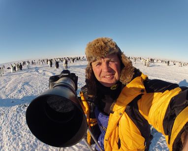 Doug Allen smiles at the camera and points his own huge camera lens at the photographer. He is at an emperor penguin colony and a hundred plus penguins can be seen behind him. Naturally it's cold, with ice and a bright blue sky!