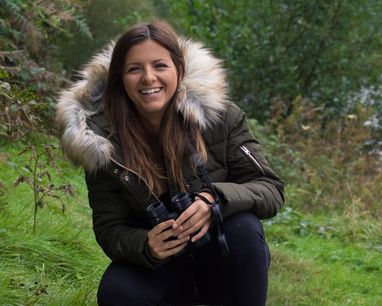 Lizzie Daly smiles at the camera against a green grass and woodland setting. She is wearing a puffer jacket with a fur hood.