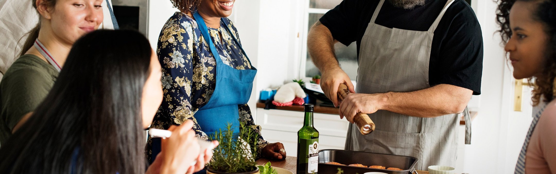 A group of adults are standing around a kitchen island, laughing and chatting as a man holding a pepper grinder puts the finishing touches to a tasty seafood meal.
