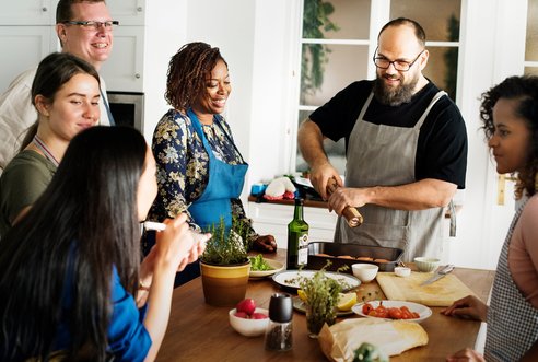 A group of adults are standing around a kitchen island, laughing and chatting as a man holding a pepper grinder puts the finishing touches to a tasty seafood meal.
