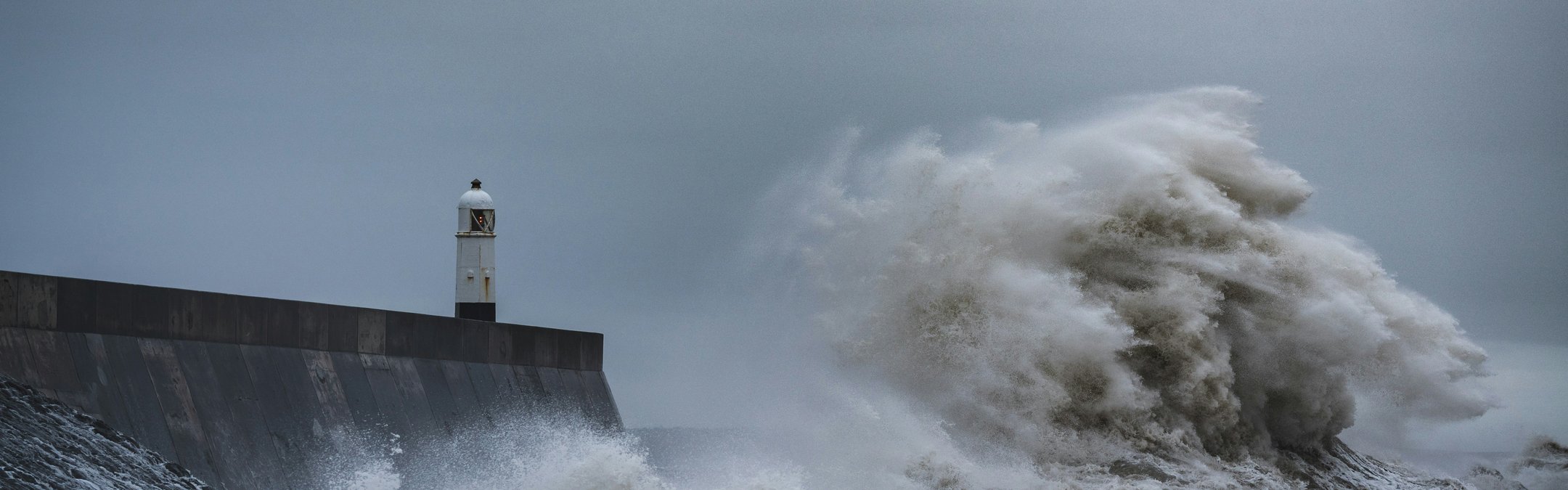 Large white waves hitting a wall at Porthcawl, Wales, with a dark grey sky above the coastline