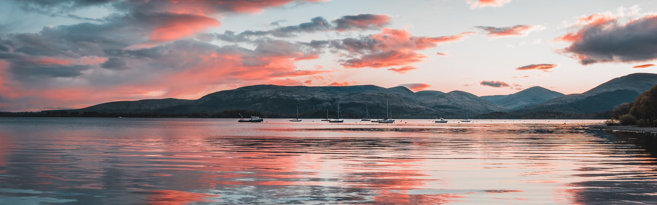 A Scottish seascape with pink and grey clouds in the sky and reflected in the water. The picture is taken from shore with land mass across the water. It looks to be dawn or dusk, with the clous hinting at either a beautiful or stormy day.
