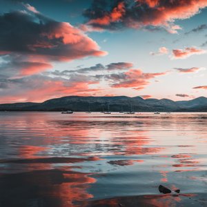A Scottish seascape with pink and grey clouds in the sky and reflected in the water. The picture is taken from shore with land mass across the water. It looks to be dawn or dusk, with the clous hinting at either a beautiful or stormy day.