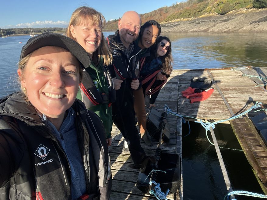 4 staff members from the Natur Am Byth porject stand huddled together on a jetty, smiling at the camera, as the sun shines behind them