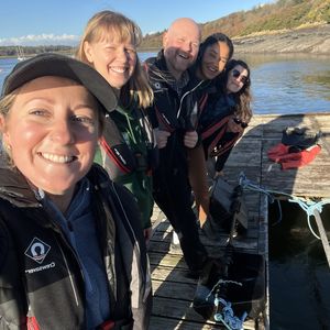 4 staff members from the Natur Am Byth porject stand huddled together on a jetty, smiling at the camera, as the sun shines behind them