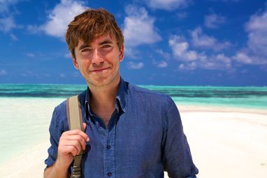 Simon Reeve stands smiling in a blue shirt against a backdrop of a white sand beach and clear blue ocean.