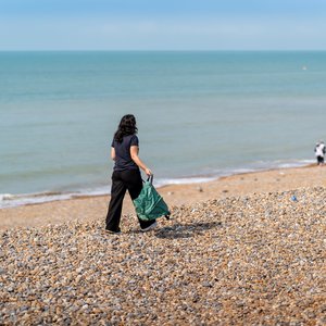 A beach cleaner walks with her back to the camera on a rocky UK beach with the sea to her left.. From the clear blue sky and her sleeveless clothing, it looks like a beautiful UK summer day.