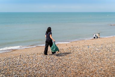 A beach cleaner walks with her back to the camera on a rocky UK beach with the sea to her left.. From the clear blue sky and her sleeveless clothing, it looks like a beautiful UK summer day.