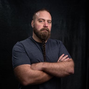 Tom 'The Blowfish' Hird looks at the camera without smiling. He has his arms crossed against a dark backdrop. His long beard is braided and his muscular arms can be seen as he is wearing a grey tshirt.