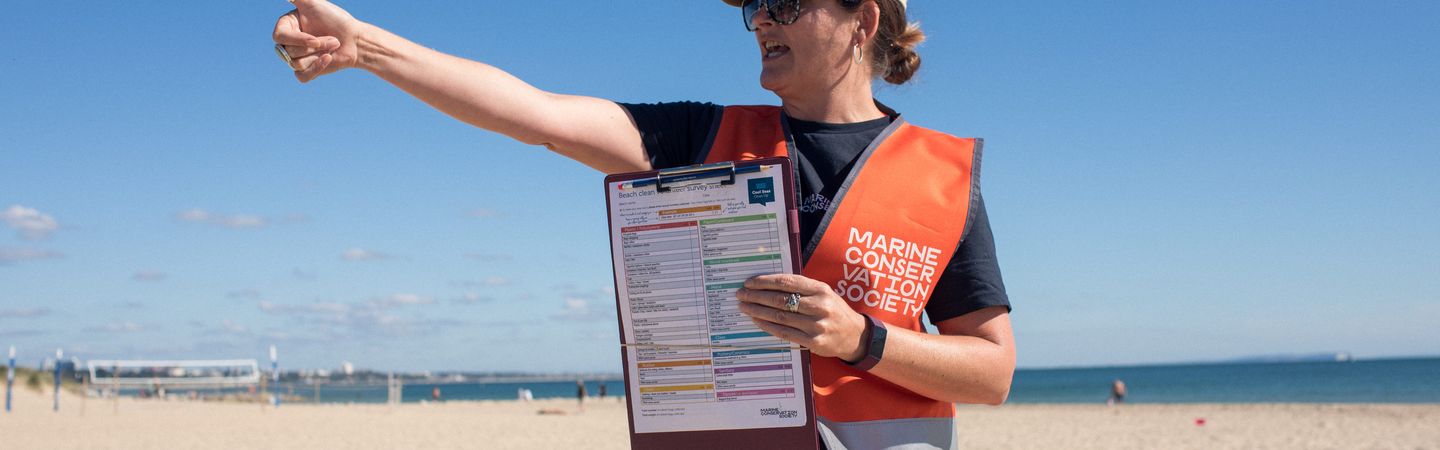 A woman with a hat and sunglasses standing on a beach. She is pointing to the left while holding a survey form for others to see. She is wearing a bright orange safety vest with a Marine Conservation society logo on it.