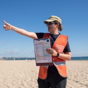A woman with a hat and sunglasses standing on a beach. She is pointing to the left while holding a survey form for others to see. She is wearing a bright orange safety vest with a Marine Conservation society logo on it.