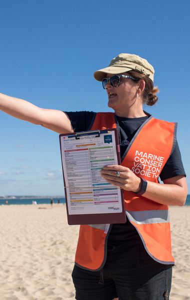 A woman with a hat and sunglasses standing on a beach. She is pointing to the left while holding a survey form for others to see. She is wearing a bright orange safety vest with a Marine Conservation society logo on it.