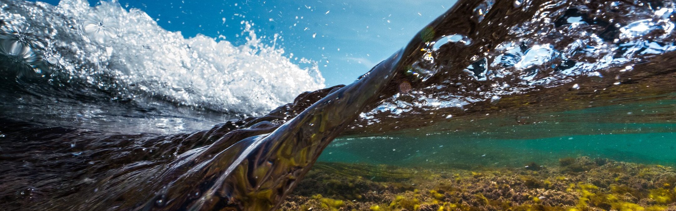 A Cornwall wave taken with the camera partially submerged in the water. The shallow water shows a rocky sea bed with algae, while a brilliant blue sky can be seen above the crashing wave.