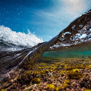 A Cornwall wave taken with the camera partially submerged in the water. The shallow water shows a rocky sea bed with algae, while a brilliant blue sky can be seen above the crashing wave.
