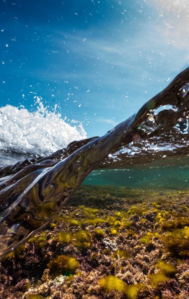 A Cornwall wave taken with the camera partially submerged in the water. The shallow water shows a rocky sea bed with algae, while a brilliant blue sky can be seen above the crashing wave.
