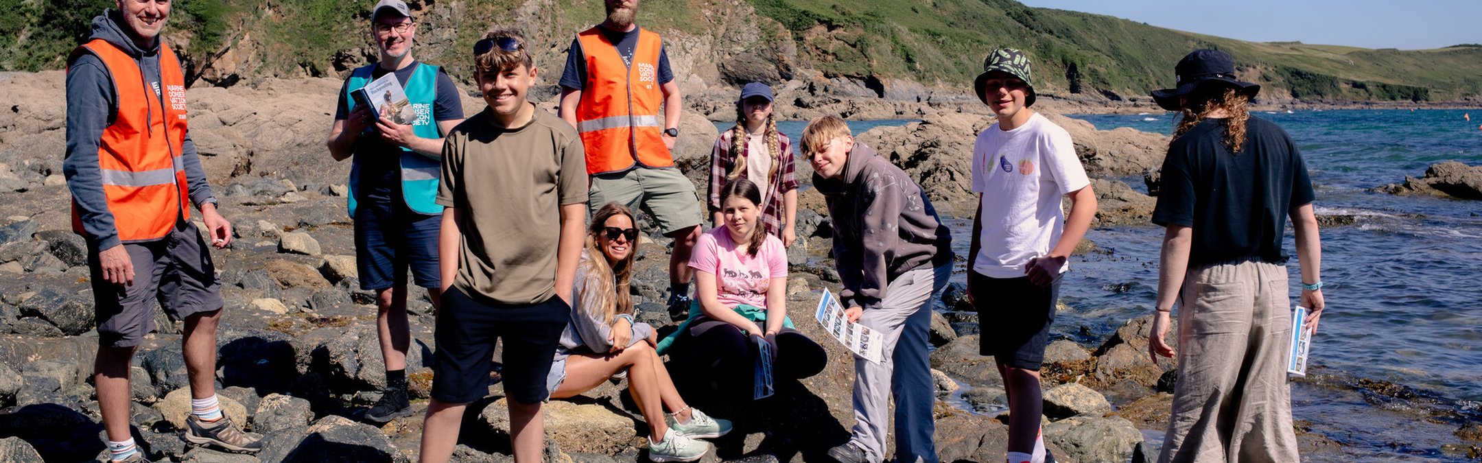 A group of young people are stood around a rockpool in the sunshine. They are with two Marine Conservation Society staff.