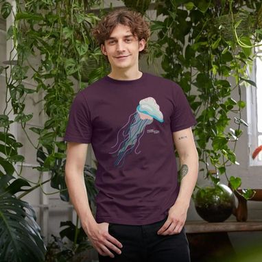 A man wearing the Marine Conservation Society's Portuguese man o'war t shirt in purple. There are green plants along the wall behind him.
