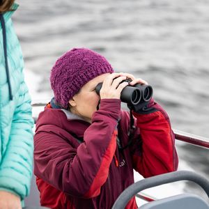 A person in a red jacket and purple beanie is using black binoculars, with another person in a teal jacket partially visible nearby. They are on a boat.