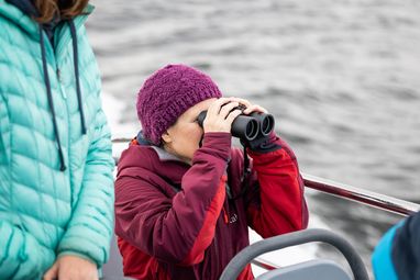 A person in a red jacket and purple beanie is using black binoculars, with another person in a teal jacket partially visible nearby. They are on a boat.