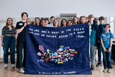 A group of young adults holding up a big tapestry with fish on it. At the top it reads "You are not a dop in the ocean you are an entire ocean in a drop - Rumi". At the bottom it reads "#Youth Ocean Network"