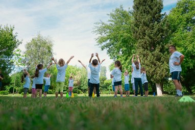 Children playing in a park surrounded by trees and grass. 2 adults can be seen with them