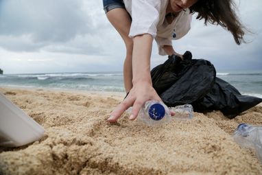 A female in a white top is reaching down to pick up a plastic bottle from the sand