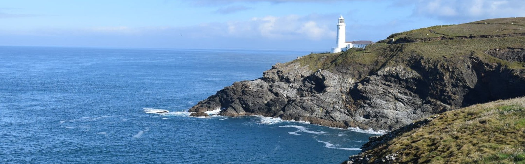 A white lighthouse standing on a rugged, grassy headland extending into the blue sea under a partly cloudy sky. The lighthouse is Trevose Head Lighthouse in Cornwall.