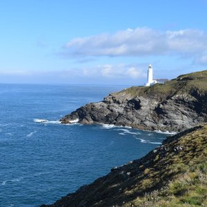 A white lighthouse standing on a rugged, grassy headland extending into the blue sea under a partly cloudy sky. The lighthouse is Trevose Head Lighthouse in Cornwall.