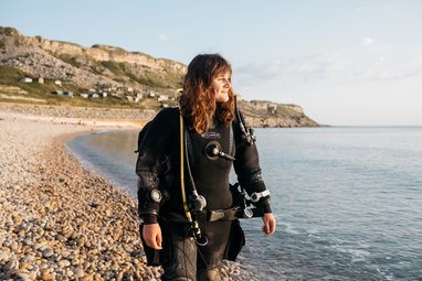 A woman wearing full black scuba diving gear, including a drysuit and a regulator, standing on a pebble beach at the water's edge, smiling and looking out towards the sea and a large cliff face in the background.