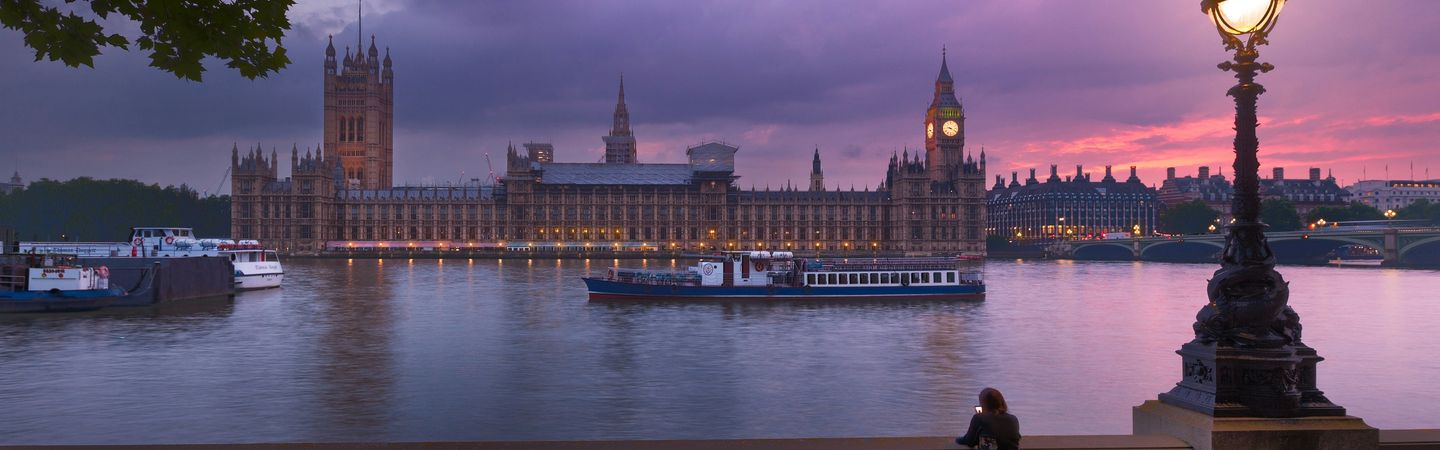 Westminster at sunset taken from the bank across the Thames. The sky is a stunning purple pink and an ornate lamp is lit in the foreground.