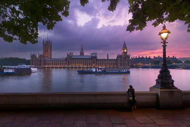 Westminster at sunset taken from the bank across the Thames. The sky is a stunning purple pink and an ornate lamp is lit in the foreground.