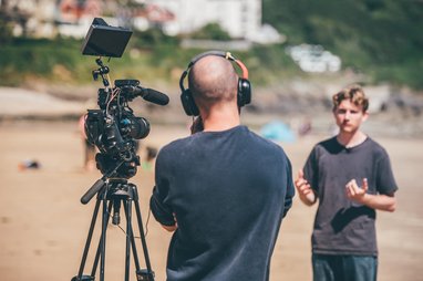 A videographer wearing headphones is operating a professional camera on a tripod on a sandy beach, while a young man stands nearby talking. The young man is blurred.
