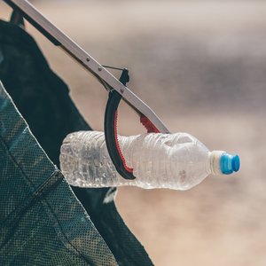 A close-up of a person using a litter picker tool to place a clear plastic water bottle with a blue cap into a dark green reusable bag. The person is out of shot.
