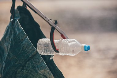 A close-up of a person using a litter picker tool to place a clear plastic water bottle with a blue cap into a dark green reusable bag. The person is out of shot.
