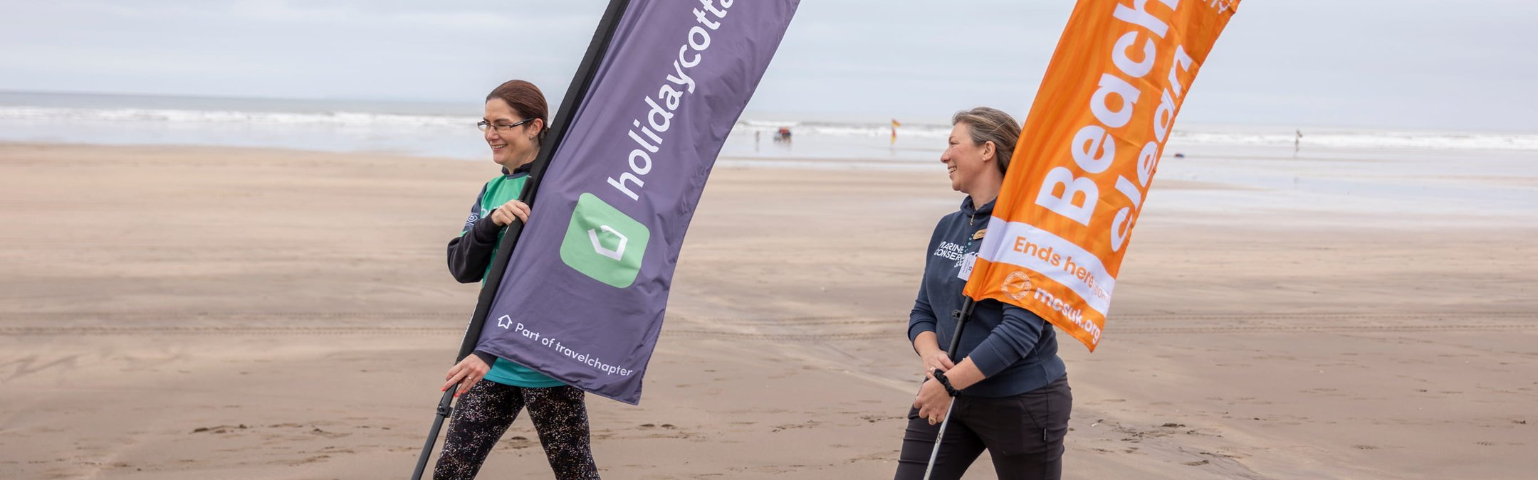 2 women walking along the beach carrying large marker flags. The flags read "Holidaycottages.co.uk" and "Marine Conservation Society Beach Clean" respectively