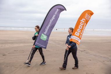 2 women walking along the beach carrying large marker flags. The flags read "Holidaycottages.co.uk" and "Marine Conservation Society Beach Clean" respectively