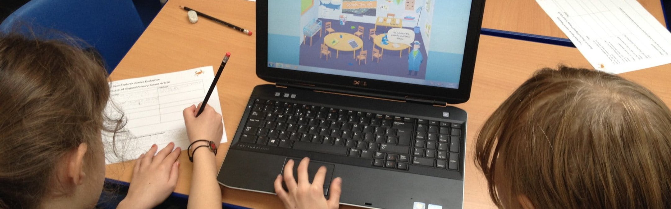 The back of 2 primary school girls' heads looking at a laptop screen on a table. The laptop screen shows a cartoon sea captain standing iinside a classroom.