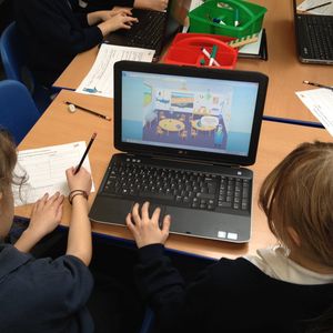 The back of 2 primary school girls' heads looking at a laptop screen on a table. The laptop screen shows a cartoon sea captain standing iinside a classroom.