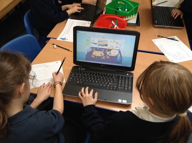 The back of 2 primary school girls' heads looking at a laptop screen on a table. The laptop screen shows a cartoon sea captain standing iinside a classroom.