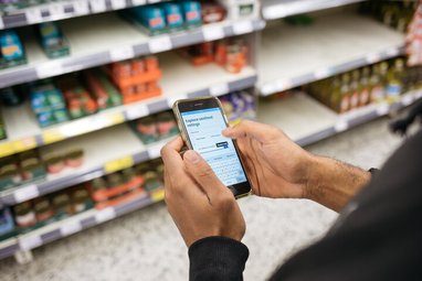 A person stands in front of the fish section in a supermarket holding a phone in their hand. On the phone screen is the Good Fish Guide species ratings page.