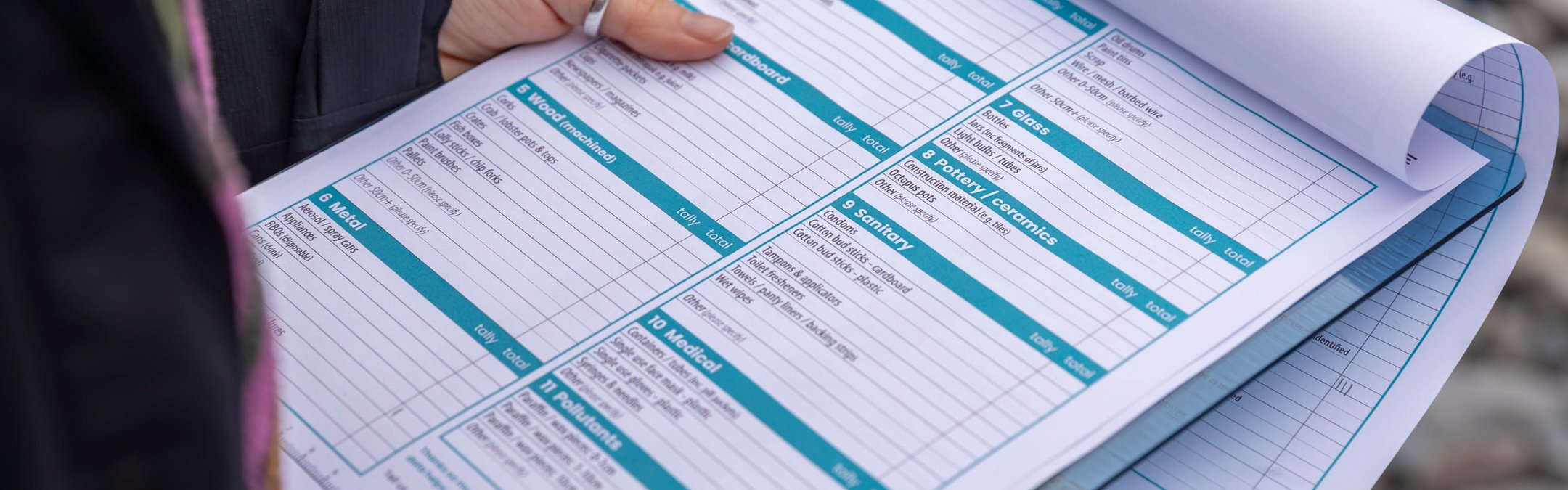A close-up shot of a person holding a clipboard with a Marine Conservation Society beach clean survey form. The user's hands and a pen are visible as they potentially make notes on the form.
