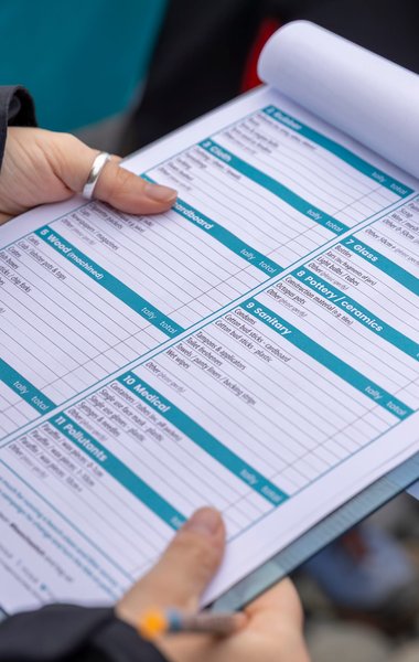 A close-up shot of a person holding a clipboard with a Marine Conservation Society beach clean survey form. The user's hands and a pen are visible as they potentially make notes on the form.