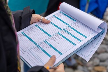 A close-up shot of a person holding a clipboard with a Marine Conservation Society beach clean survey form. The user's hands and a pen are visible as they potentially make notes on the form.