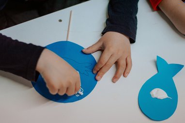 the hands of a child making a fish out of blue paper, glue and a stick