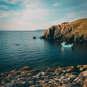 A small fishing boat is pictured leaving Abercastle Harbour. The water is a deep blue and there are craggy rocks to the right of the photo.