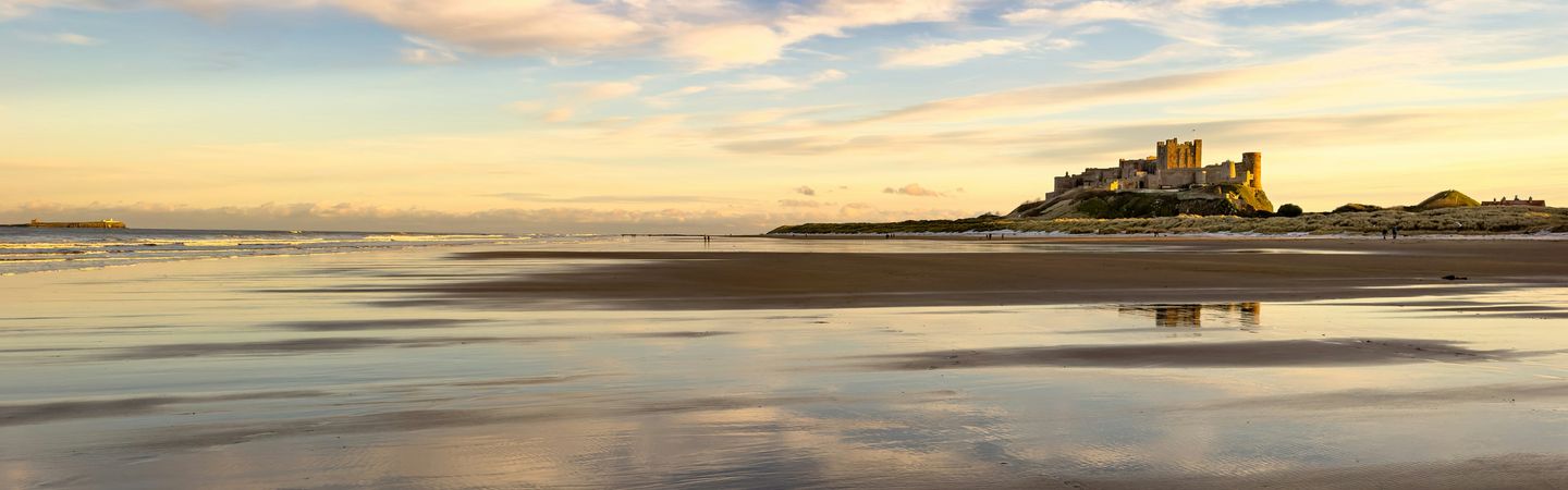 Bamburgh Castle as seen across Bamburgh Castle Beach. It looks like dusk, with a pale golden sky on the horizon and the colours reflecting off the water on the sand.